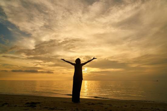 A silhouette of  a woman raising her hands in prayer by the seashore at sunset.