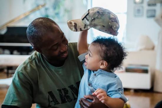 A father in a green army shirt smiling at a crying baby boy to calm him.