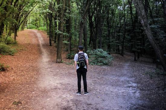 A man carrying a backback stranded at a crossroad in the woods wondering what path to take.