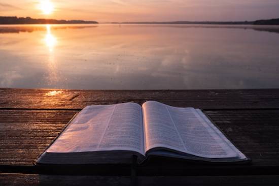 An Open Bible An open Bible on a Table close to a waterbody reflecting on the rays of the setting sun.