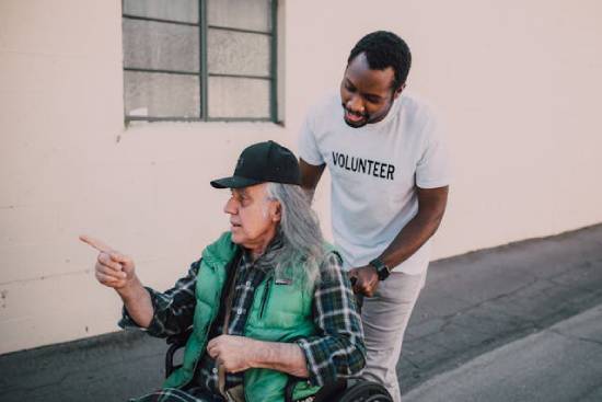 A Volunteer Caregiver A gentleman in white T-shirt volunteering to help move an old lady with her wheelchair.