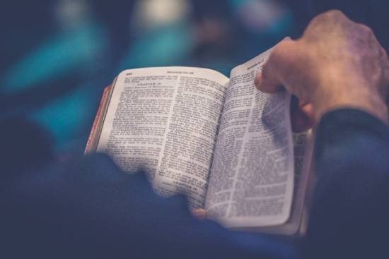 A close-up view of a person's hand holding the page of an open Bible.