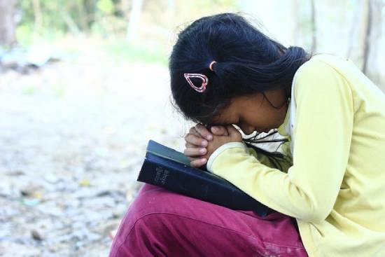 Praying Kid A kid in a mustard jumper and maroon trouser praying with a bible on her kness.