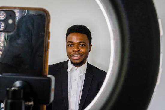 A man in a black blazer and a white shirt standing infront of a camera vlogging.