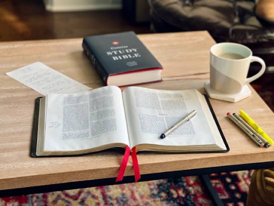 Bible study tools on table including an open bible, closed bible, writing leaflet, pens, and a cup of beverage.